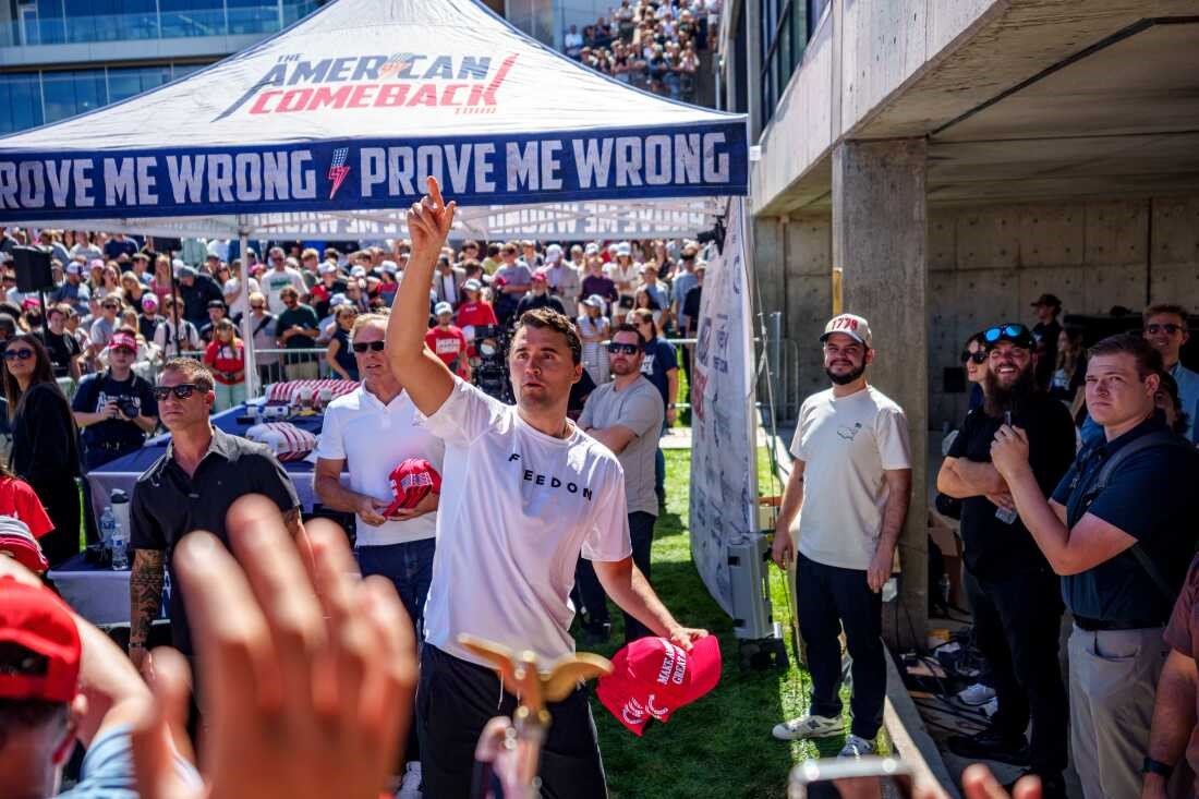 Charlie Kirk at a rally handing out red hats with American Comeback tent in background