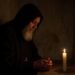Saint Charbel Makhlouf in prayer wearing a black Maronite hood and robe in a dark candlelit monastery